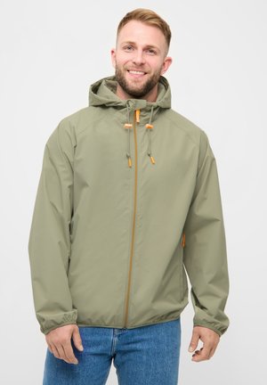 Smilende mand iført en lys grøn hættetrøje med orange lynlås og snore, kombineret med blå jeans mod en ensfarvet baggrund.