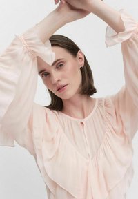 Vaaleanpunainen pusero, jossa on kerroksellisia rypytyksiä vartalon ja hihojen ympärillä. Pehmeä, läpikuultava kangas, jossa on napitus edessä ja leveät hihansuut.