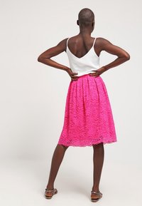 Kobieta stoi z rękami na biodrach, w białym bezrękawniku, jasnoróżowej koronkowej spódnicy i brązowych sandałach, zwrócona plecami w stronę gładkiego tła.