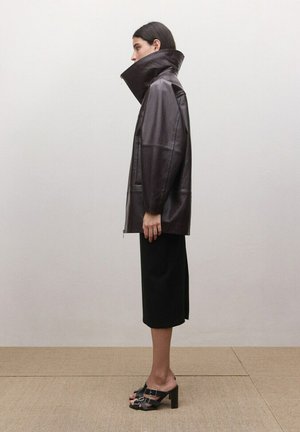 Mujer de perfil con una chaqueta de cuero marrón oscuro sobredimensionada con cuello alto, falda midi negra y sandalias negras de tacón, de pie sobre un suelo beige.