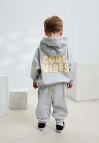 Hallikas kapuutsiga pusa, mille tagaosas on suur beež "COOL VIBES" tekst, mida kannab seljas sobivate hallide dressipükste ja mustade tennistega komplekt. Pehme kangas.