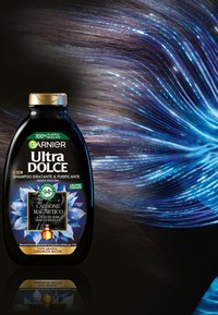 La bottiglia dello shampoo Garnier Ultra Dolce è nera con un tappo dorato. Presenta accenti floreali blu e testi riguardanti ingredienti per l'idratazione e la purificazione.