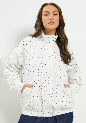 Frau mit langen braunen Haaren, die eine weiße Jacke mit schwarzen Punkten trägt, Hände in den Taschen, stehend vor einem einfarbigen hellen Hintergrund.