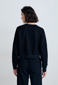 Svart cropped hettegenser med teksturert geometrisk mønster, ribbede ermer og nederkant, samt rund halslinje. Bæres over matchende bukser.
