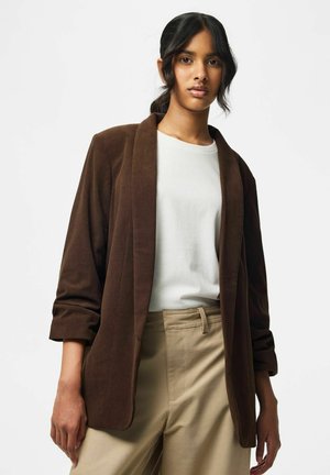 Giovane donna con capelli scuri che indossa un blazer in velluto a costine marrone, una maglietta bianca e pantaloni beige su uno sfondo chiaro uniforme.