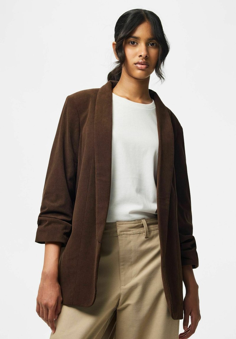 Jeune femme aux cheveux foncés portant un blazer en velours côtelé marron, un haut blanc et un pantalon beige devant un fond clair uni.