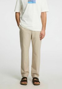 Beige bomullsblandade byxor med en avslappnad passform, slät textur och subtila veckdetaljer. Kombinationen med en vit t-shirt med grafisk tryck och mörka sandaler.