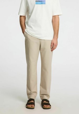 Pantaloni in misto cotone beige con vestibilità rilassata, texture morbida e dettagli di piega sottili. Abbinati a una maglietta bianca con grafica e sandali scuri.