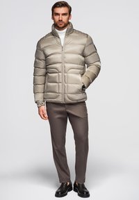 Puffet lys beige jakke med høj krave, vandret syninger og sidelommer, parret med brune bukser og sorte sko.