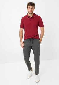 Polo rouge avec un col contrasté, associé à un pantalon gris avec cordon de serrage et des baskets blanches, se tenant contre un fond blanc.