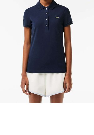 Osoba ubrana w granatową koszulkę polo z krótkim rękawem z białymi guzikami i białe szorty na jednolitym jasnym tle.