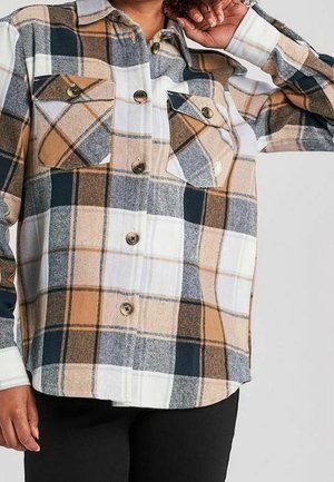 Person trägt ein beige-weiß-grau-schwarz kariertes Hemd mit Knöpfen und zwei Brusttaschen, von denen eine teilweise aufgeknöpft ist, sowie schwarze Hose.