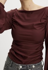 Maglietta bordeaux a maniche lunghe con scollatura a barca e dettagli arricciati sulle spalle, abbinata a jeans neri a vita alta con texture.