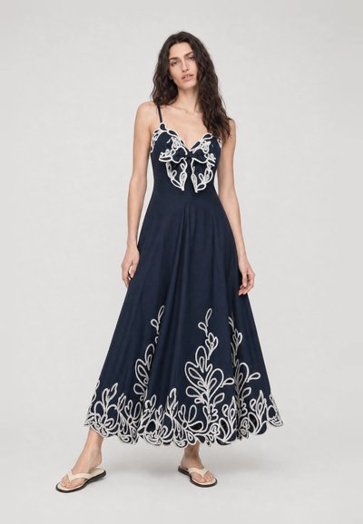 Femme portant une robe bleu marine sans manches avec broderie florale blanche et détail nœud, associée à des sandales plates beige, debout sur un fond blanc.