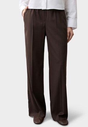 Personne portant un pantalon large marron foncé avec des plis à l'avant, une chemise blanche et des chaussures marron, debout avec une main dans la poche.