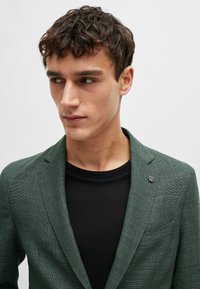 Blazer verde strutturato con una vestibilità slim, rever a punta e un piccolo dettaglio a spilla. Indossato sopra una t-shirt nera a fantasia.