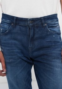 Jeans din denim albastru închis, cu un design clasic cu cinci buzunare, având detalii uzate și un închizător cu buton la talie.