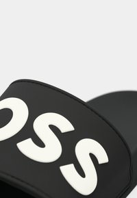 Infradito nera con una fascia ampia caratterizzata da lettere bianche in rilievo. Materiale sintetico liscio con punta arrotondata e design minimalista.