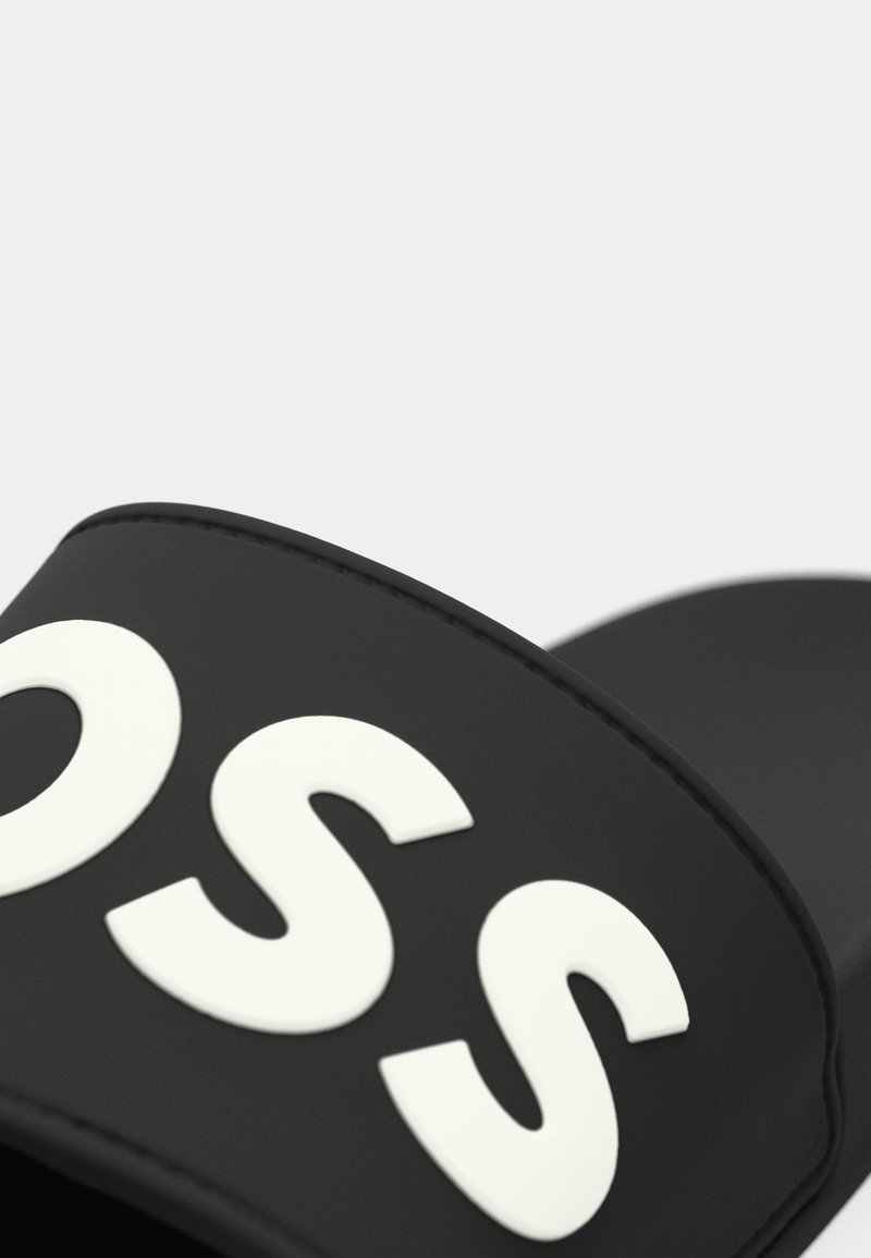 Schwarze Slidesandale mit einem breiten Riemen, der erhabene weiße Buchstaben zeigt. Glattes synthetisches Material mit runder Zehenpartie und einem minimalistischen Design.