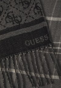 Écharpe tissée noire et grise présentant un motif géométrique, avec des bords à franges et un détail de logo subtil en gris, fabriquée en tissu doux.