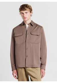 Hellbraunes Button-Up-Hemd mit zwei Brusttaschen, langen Ärmeln, spitzem Kragen und glatter Stoffoberfläche. Über einem weißen Oberteil getragen.
