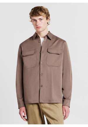 Camicia button-up color marrone chiaro con due tasche sul petto, maniche lunghe, colletto a punta e texture del tessuto liscia. Indossata sopra un top bianco.