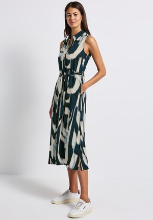 Robe sans manches, ceinturée, dans un motif abstrait vert foncé et crème. Comprend un col, une fermeture à boutons et des poches latérales. Associée à des baskets blanches.