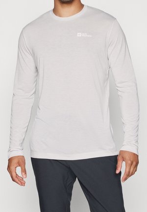 Man draagt een lichtgrijs Jack Wolfskin shirt met lange mouwen en donkere broek, staat met ontspannen armen langs zijn zij.