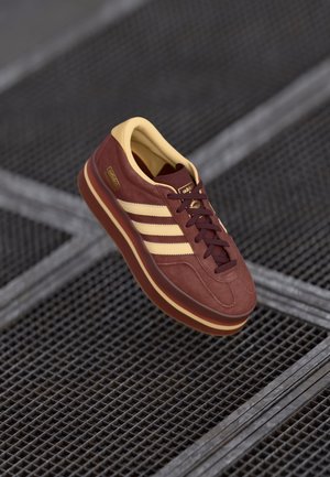 Baskets Adidas marron et beige avec trois bandes flottant au-dessus d'une surface en grille métallique noire.
