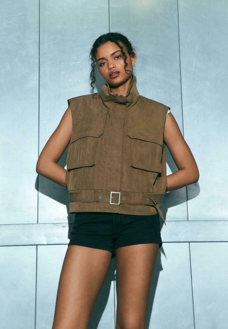 Donna con capelli ricci che indossa un gilet marrone senza maniche e pantaloncini neri, in piedi davanti a uno sfondo metallico con le mani dietro la schiena.