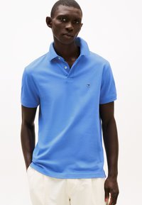 Polokošile s krátkým rukávem v jasně modré bavlně s dvouknopovou légou a malou sadačkou s logem na levé straně hrudi. Hladká textura.