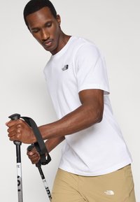 Λευκό βαμβακερό μπλουζάκι με κοντά μανίκια και μαύρο λογότυπο στο αριστερό στήθος. Το μοντέλο κρατάει μπαστούνια πεζοπορίας και φοράει μπεζ σορτς.