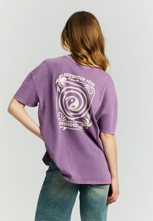 Frau mit langen Haaren trägt ein lockeres lila T-Shirt mit weißer Spirale und dem Text "Saltwater Soul Balanced All" auf der Rückseite, kombiniert mit blauen Jeans.