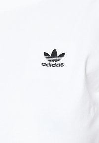 T-shirt en coton blanc avec un petit logo Adidas brodé en noir, comportant un design en trèfle et du texte en dessous. Texture lisse, encolure ronde.
