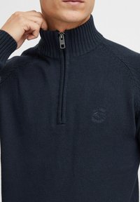 Pull en tricot marine avec col haut et fermeture éclair deux tiers. Présente une texture côtelée et un logo en relief subtil sur la poitrine.