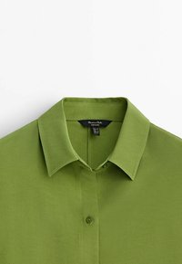 Chemise à col vert en tissu lisse, dotée d'une patte de boutonnage et d'une petite étiquette à l'intérieur du col.