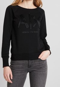 Felpa nera con colletto a girocollo e polsini a costine, caratterizzata da un logo 'AIX' testurizzato e il testo 'Armani Exchange' al centro.