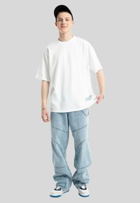 Biała, oversize'owa koszulka z niebieskimi haftami kwiatowymi, w zestawie z jasnoniebieskimi, luźnymi dżinsami z zakrzywionymi szwami oraz czarno-białymi trampkami.