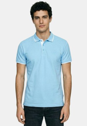 Jeune homme portant un polo à manches courtes bleu clair avec des bordures blanches, debout devant un fond clair uni.