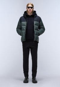 Groene en zwarte pufferjack met een capuchon, zwart sweatshirt eronder, zwarte broek en zwarte schoeisel. Vriendelijke textuur, strakke pasvorm.