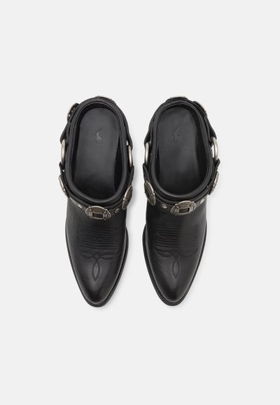 Zapatos de cuero negro hasta el tobillo con punta afilada, que cuentan con una correa decorativa y acentos de hebilla metálica, textura suave y diseño grabado.