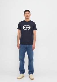 Tricou din bumbac bleumarin cu un logo grafic alb, asortat cu blugi albaștri și adidași bej; modelul stând în fața unui background neutru.