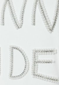 Weißer Stoff mit erhabenen Buchstaben, verziert mit weißen Perlen und klaren Strasssteinen, die ein strukturiertes Detail im gesamten Design erzeugen.
