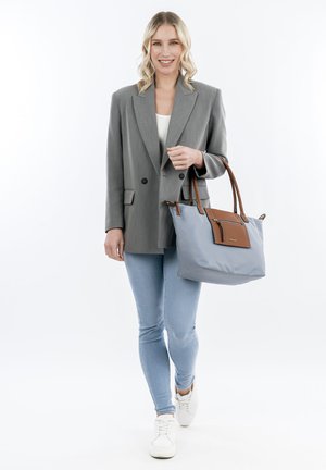 Sac en toile bleu clair avec des accents en cuir brun, poche zippée et bretelles, associé à un blazer gris et un jean clair.