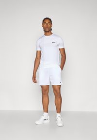 Valkoinen lyhythihainen urheilupaita, jossa vaakasuorat viivat ja 'BOSS'-logo; yhdistetty valkoisiin shortseihin ja valkoisiin tennareihin.