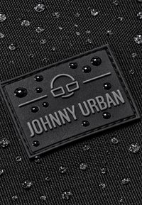 Tessuto nero testurizzato con gocce d'acqua e una toppa rettangolare nera con la scritta "Johnny Urban" con un semplice logo di occhiali sopra.