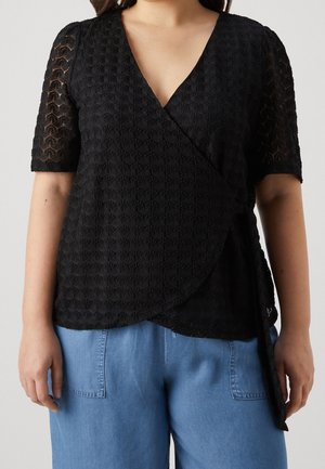 Femme portant un haut noir texturé à manches courtes et un pantalon bleu, sur un fond gris uni.