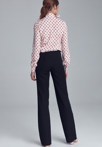 Kobieta stojąca plecami, ubrana w białą bluzkę w czerwone kropki i czarne spodnie o wysokim stanie, na jednolitym szarym tle.