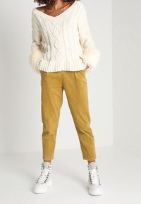 Pull crème en tricot torsadé avec poignets en fourrure, associé à un pantalon en velours côtelé moutarde et des baskets montantes blanches.