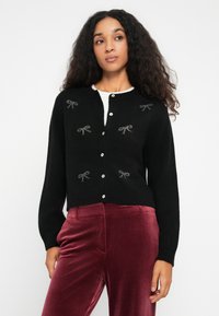 Femeie îmbrăcată într-un cardigan negru cu nasturi, decorat cu modele mici de fundițe, combinat cu pantaloni din catifea bordeaux, cu talie înaltă, pe un fundal alb.